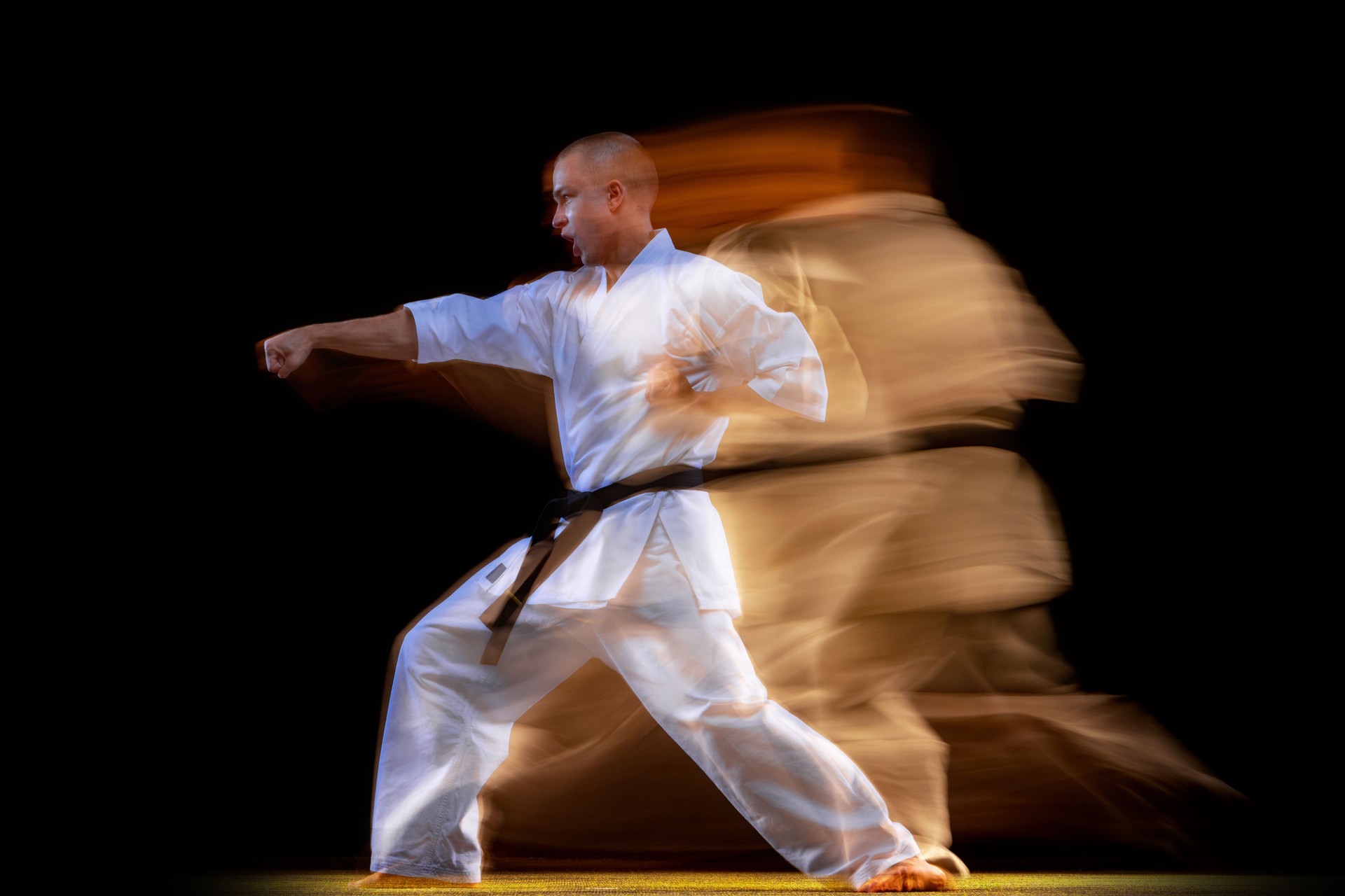 Full-length portrait of young karate sportsman wearing white kimono isolated over black background. Mixed light effect