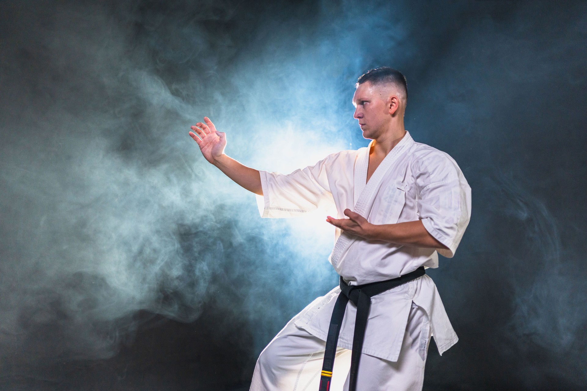 Strong man fighter in white kimono for karate martial arts with black belt preparing for fight training in black background. Man training moves punches kicks practicing shadow fight ready for sparring.